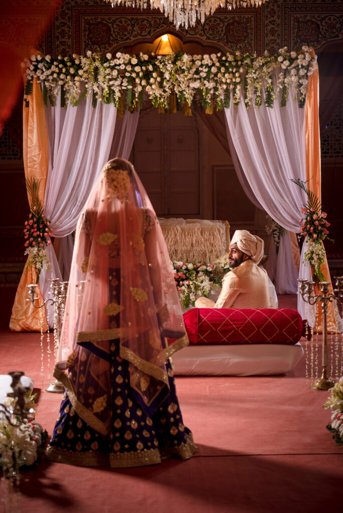Sikh wedding ceremony with bride approaching groom under floral mandap with draped decor and warm lighting